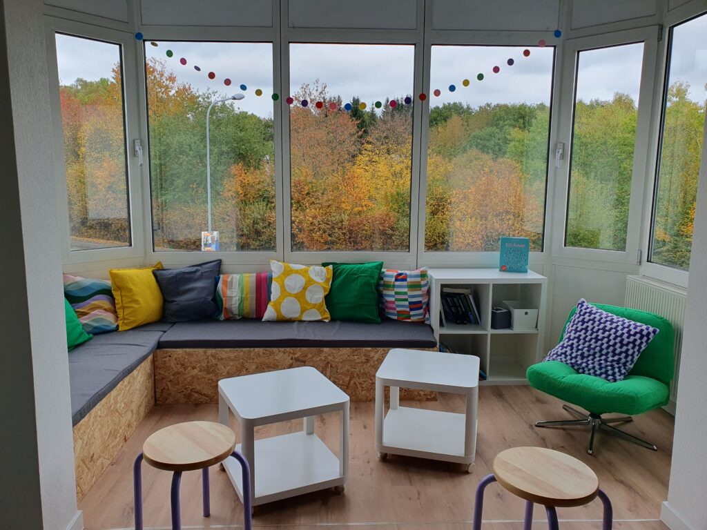 Eine gemütliche Ecke mit bunten Kissen, zwei kleinen Tischen, einem grünen Stuhl und herbstlichen Bäumen, die durch die großen Fenster zu sehen sind; geschmückt mit einer Girlande aus Filzkugeln.