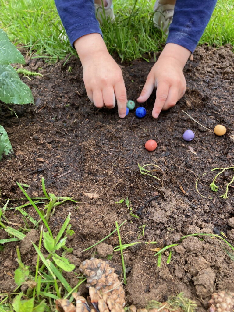 Die Hände eines Kindes legen bunte Murmeln in die Erde im Freien, umgeben von Gras und Pflanzen.