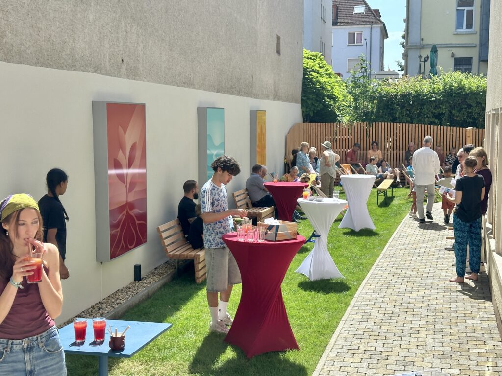 Die Leute versammeln sich in einem sonnigen, schmalen Innenhof, wo Getränke serviert werden, Kunstwerke an den Wänden hängen und Tische mit rot-weißen Tischdecken stehen. Einige sitzen auf Bänken, während andere stehen und sich unterhalten.