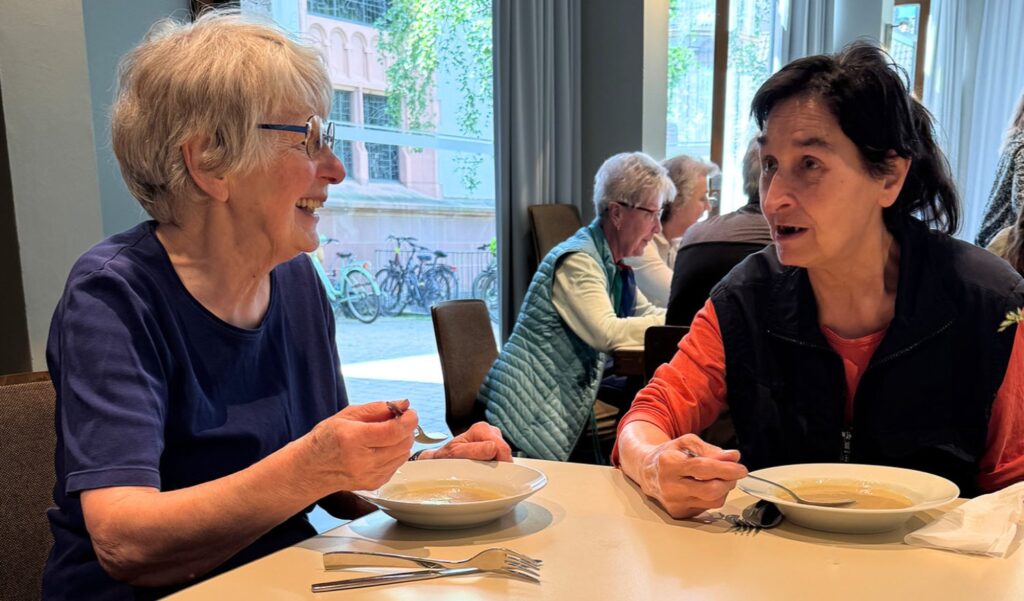 Zwei ältere Frauen sitzen an einem Tisch, lächeln und essen gemeinsam Suppe in einem hellen, ungezwungenen Speiseraum, im Hintergrund sind weitere Personen zu sehen.