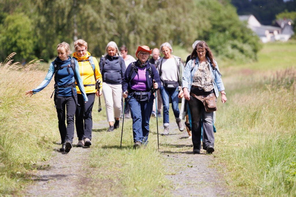 Eine Gruppe von Menschen geht im Freien auf einem grasbewachsenen Weg spazieren, umgeben von Bäumen; sie tragen Jacken und haben Rucksäcke dabei.