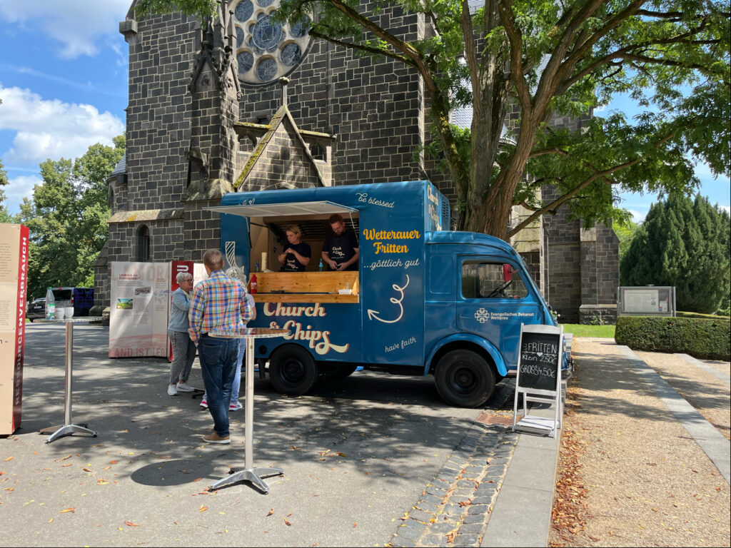 Ein blauer Imbisswagen mit der Aufschrift „Church & Chips“ steht in der Nähe einer Steinkirche und bedient an einem sonnigen Tag seine Gäste. Drei Personen stehen neben dem Wagen.