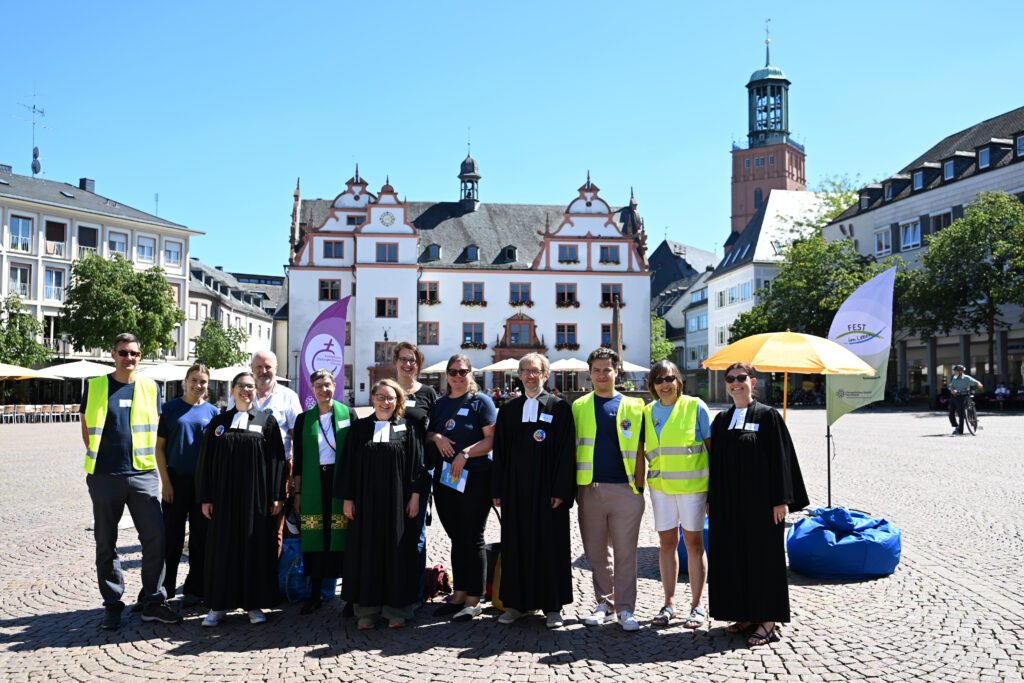Eine Gruppe von Menschen in schwarzen akademischen Roben und gelben Westen posiert gemeinsam auf einem sonnigen Stadtplatz, mit historischen Gebäuden und Bannern im Hintergrund.