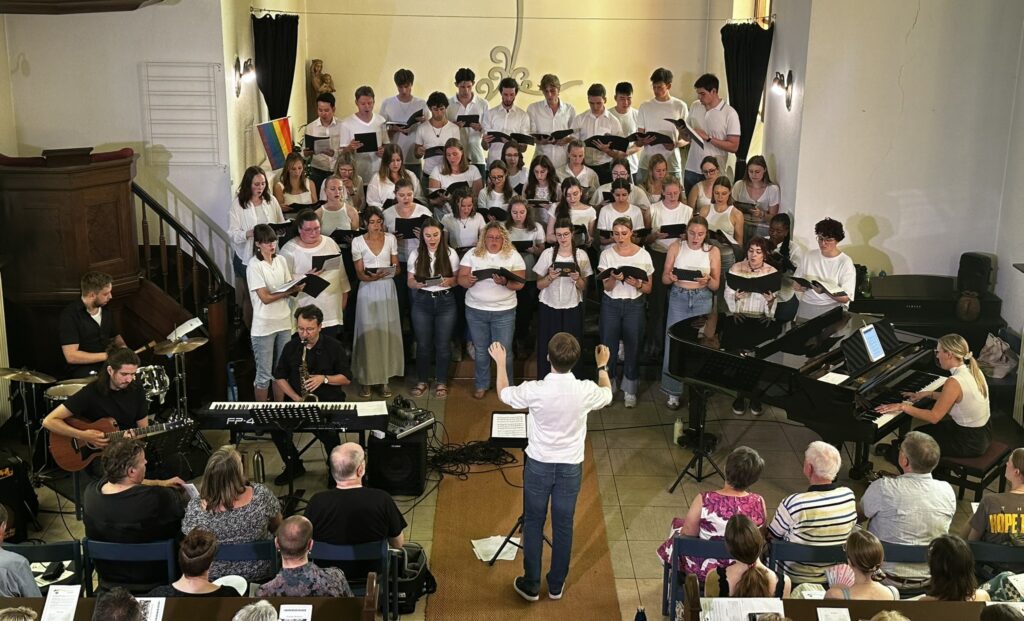 Ein Chor von Männern und Frauen in weißen Hemden singt auf der Bühne, begleitet von Musikern, während das Publikum in einer warm beleuchteten Halle zusieht.