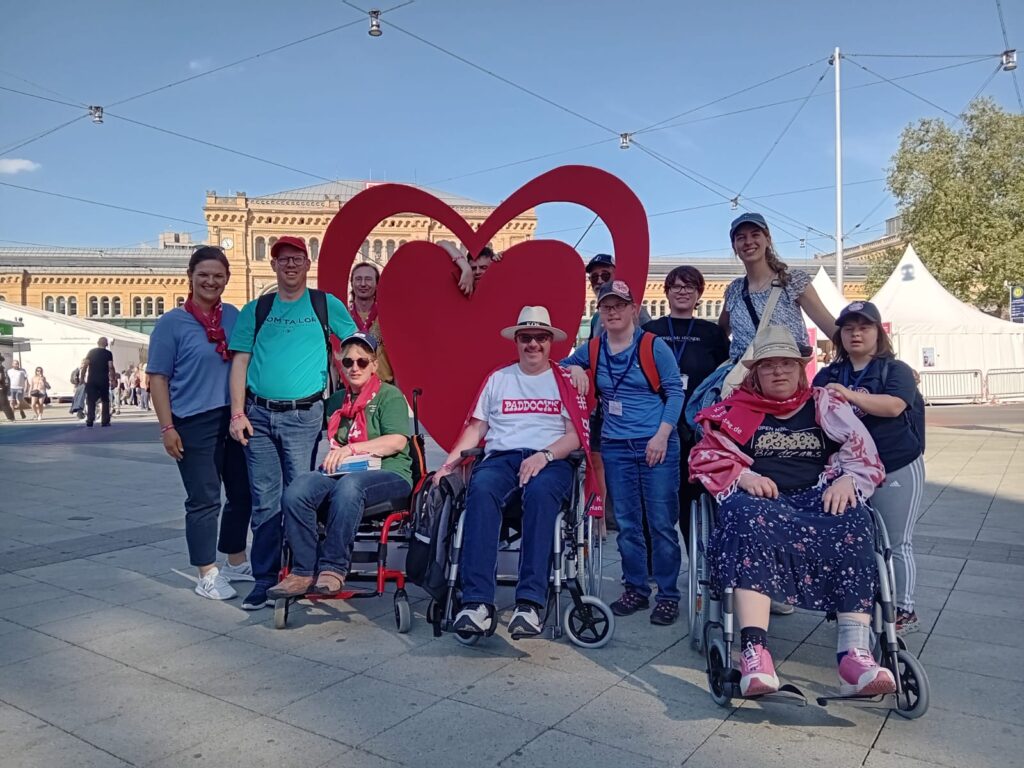 Eine Gruppe von Menschen, einige in Rollstühlen, posiert und lächelt vor einer großen roten Herzskulptur im Freien an einem sonnigen Tag.