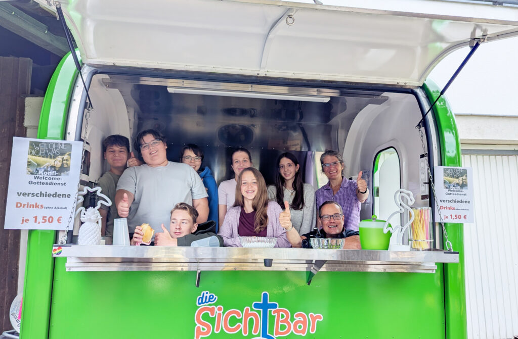 Eine Gruppe lächelnder Menschen zeigt Daumen nach oben und hält Getränke an das Fenster eines grünen Imbisswagens mit der Aufschrift die SichtBar. Zwei Schilder werben für Getränke für 1,50 €.