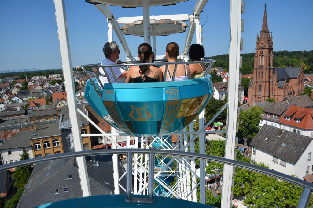 Vier Personen sitzen in einer Riesenradgondel und blicken an einem sonnigen Tag auf eine Stadt mit Häusern und einer Kirche mit einem hohen Kirchturm.