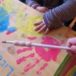 Die Hand eines Kindes ruht auf einem Tisch, der mit bunten Handabdrücken bedeckt ist, während eine andere Person einen Pinsel bereithält, um weitere Abdrücke zu machen.