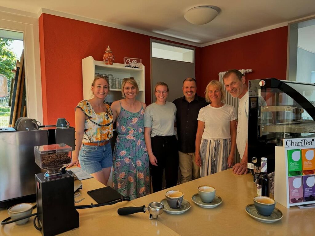 Sechs Personen stehen lächelnd hinter einem Tresen mit Kaffeetassen und -maschinen in einem gemütlichen, warm beleuchteten Café mit roten Wänden und heimeliger Einrichtung.