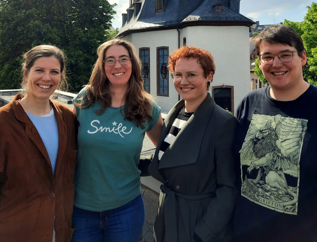 Vier lächelnde Menschen stehen gemeinsam im Freien vor einem weißen Gebäude mit Bäumen, sind leger gekleidet und genießen einen sonnigen Tag.