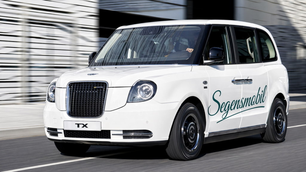 Ein weißes Taxi mit dem Schriftzug Segensmobil auf der Seite fährt eine Straße in der Stadt entlang, mit schwarzem Kühlergrill und modernem Design.