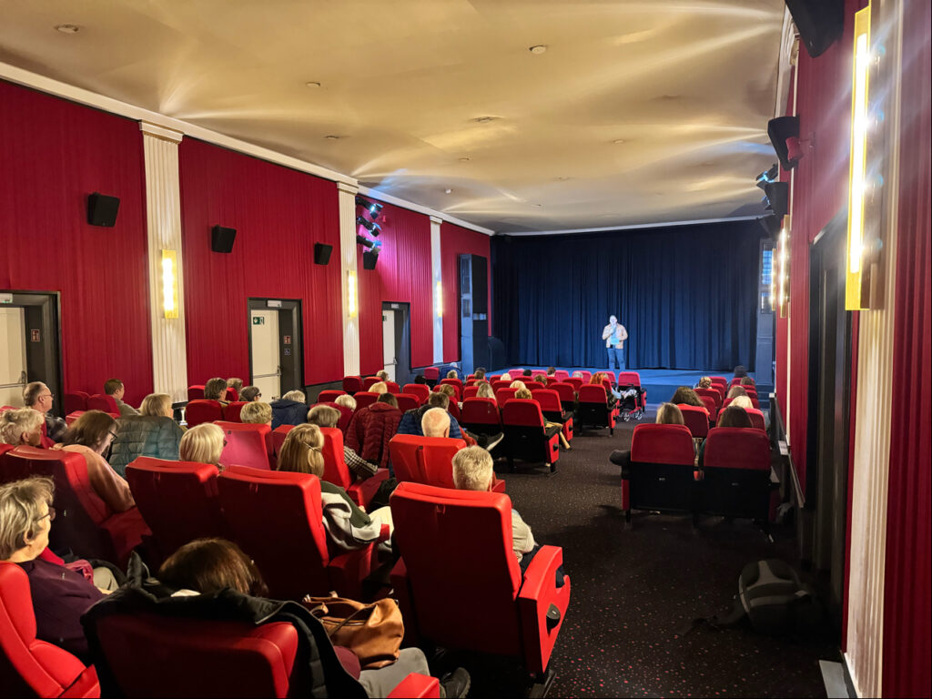 Eine Person steht auf der Bühne und spricht zu einem Publikum, das auf roten Stühlen in einem Theater mit roten Wänden und weißen Säulen sitzt.