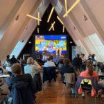 In einem modernen Konferenzraum sitzen die Teilnehmer und verfolgen zwei Referenten, die vorne auf einer großen Leinwand ihre Folien präsentieren. Der Raum hat schräge Wände und Hängeleuchten.