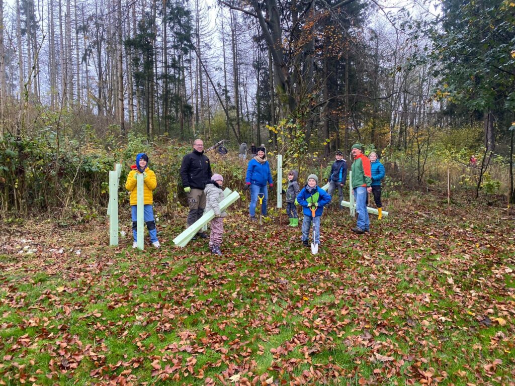 Eine Gruppe von Kindern und Erwachsenen steht in einem Waldgebiet mit Materialien zum Baumpflanzen.